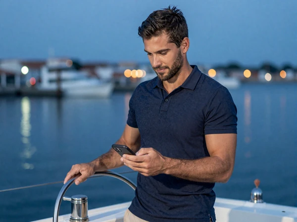 Mann in Navy-Polo am Yacht-Ruder bei D&auml;mmerung mit Smartphone - Casea Casino Sportwetten Bonus